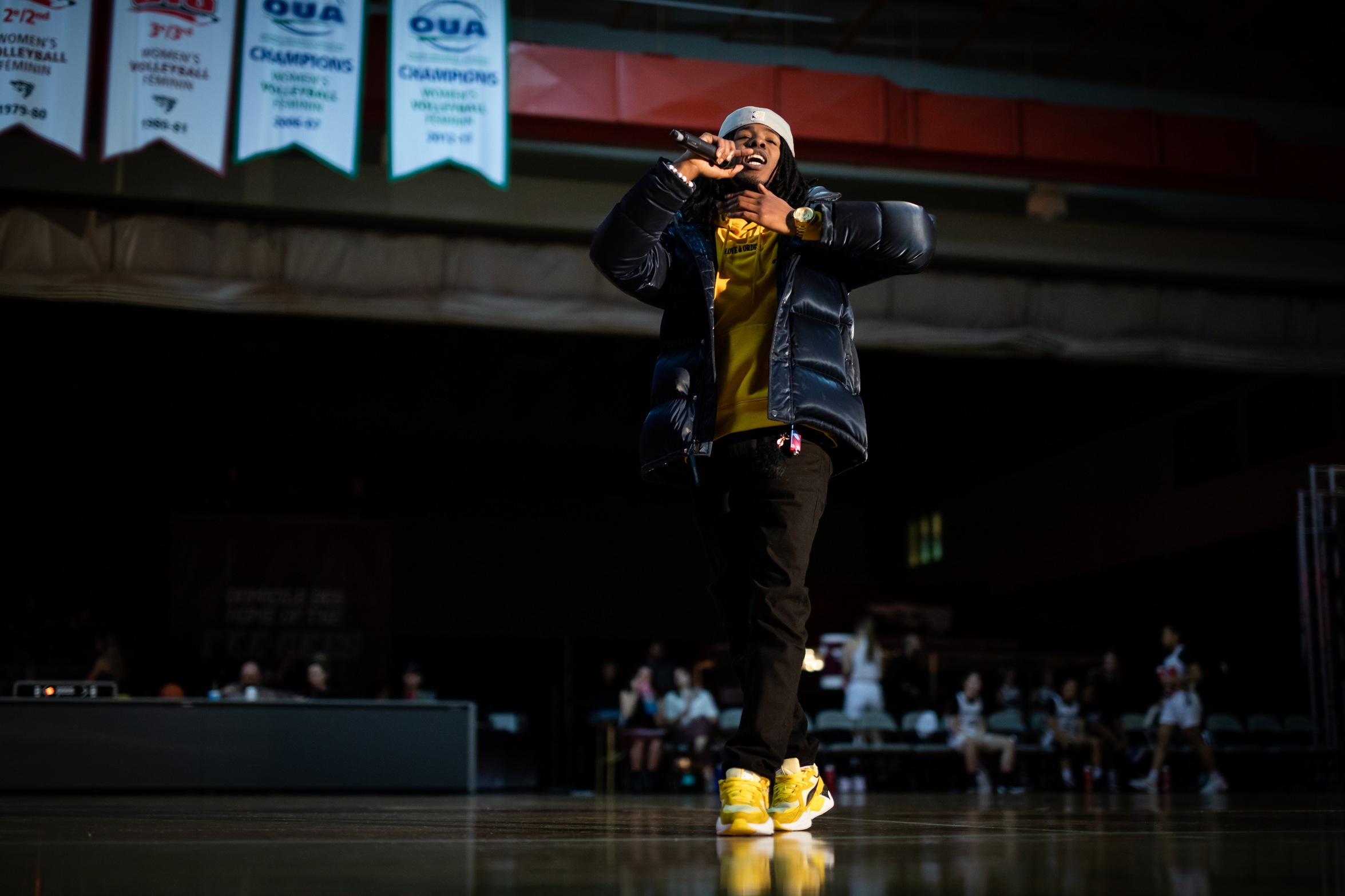 A performer with awesome yellow shoes speaks into a microphone in a dark gymnasium.