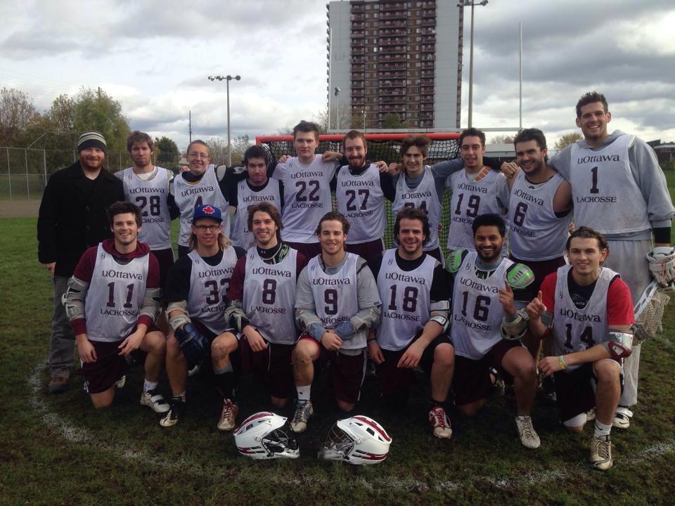 Team of men's lacrosse players pose on a grass field with an apartment building behind them