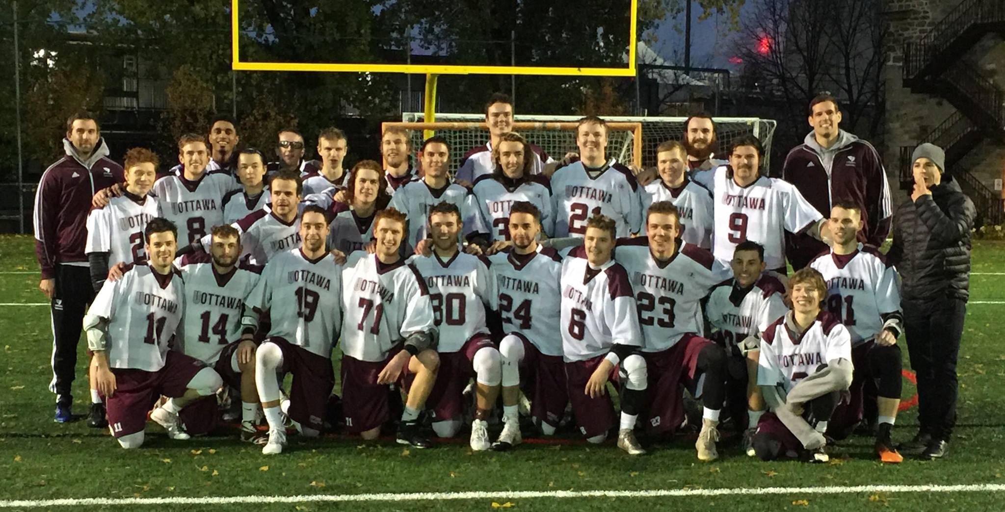 Team photo of men's lacrosse players under the football goalposts on an artificial turf field.