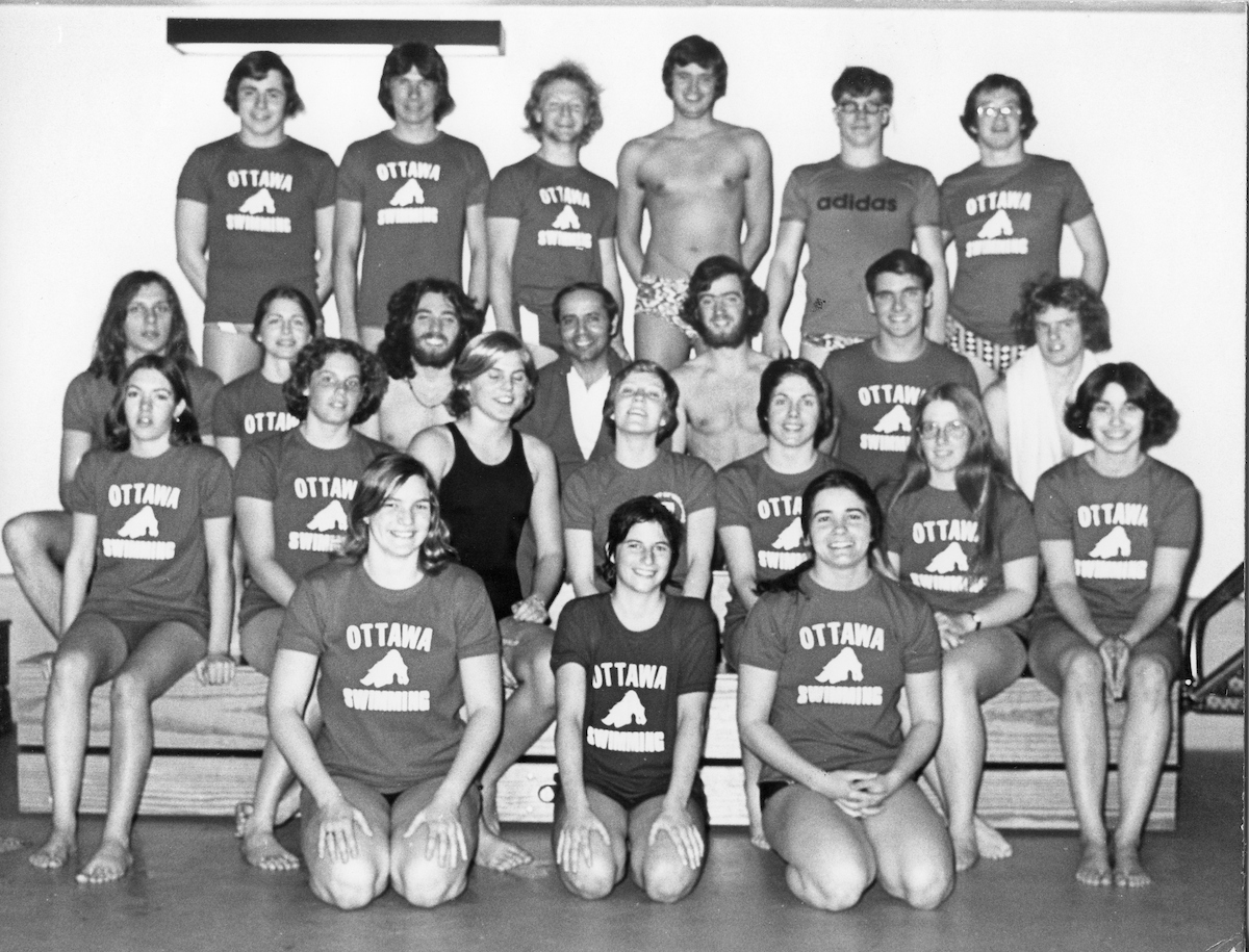 Black and white photo of the Gee-Gees swim team wearing team t-shirts, seated and standing around coach Fouad Kamal, in a dark suit, in the middle of the photo.