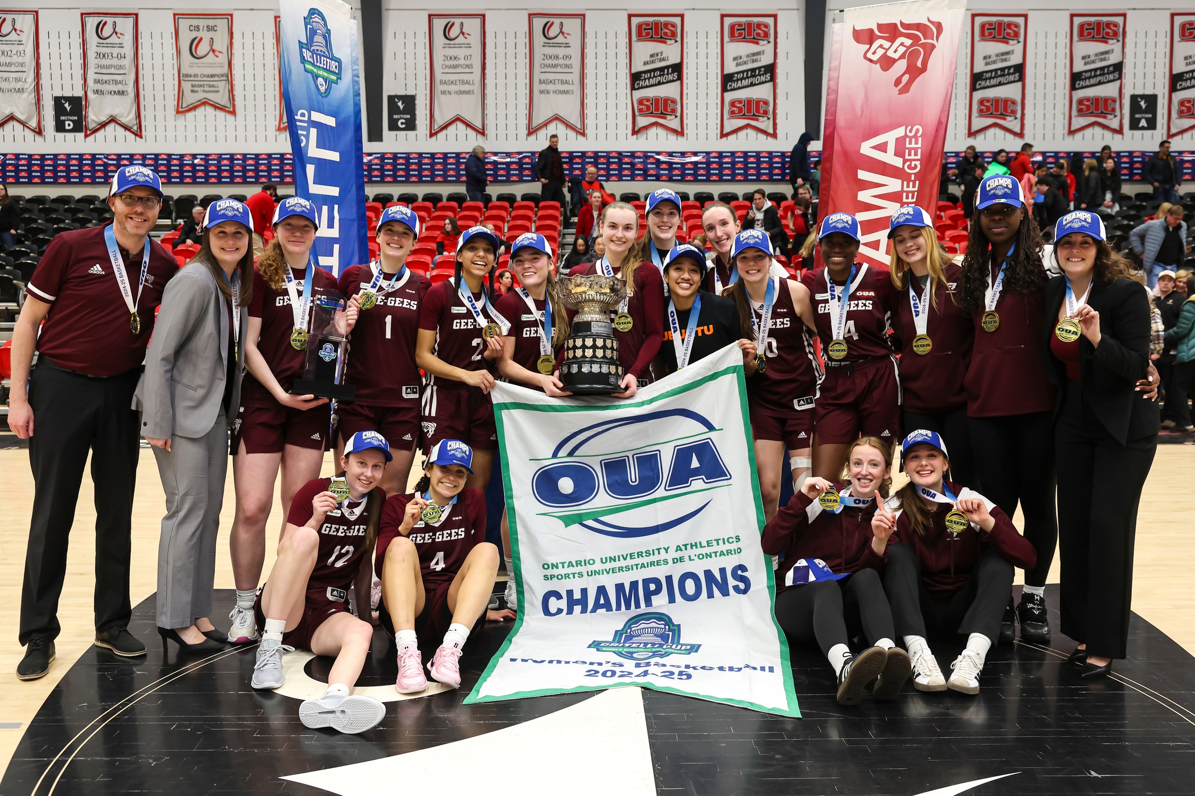 Gee-Gees women's basketball team players and coaches pose with the OUA Championship banner and Critelli Cup trophy, and their gold medals.