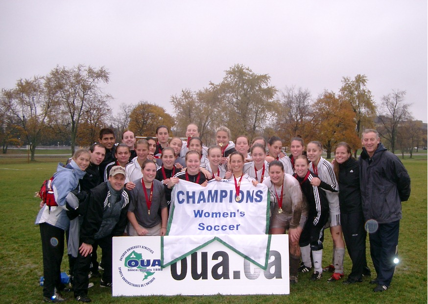 The 2003 Gee-Gees women's soccer team poses on the field with an OUA sign and the Championship Banner. The players are wearing medals with red ribbons.